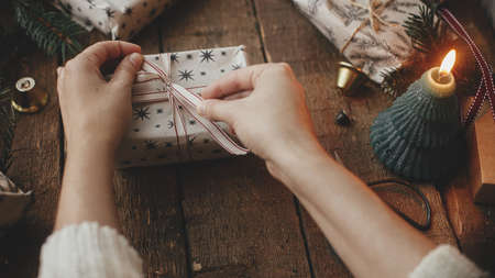 Hands wrapping stylish christmas gift in craft paper on rustic wooden table with candles, scissors, fir branches. Merry Christmas! Stylish scandinavian xmas presents, atmospheric moody imageの写真素材