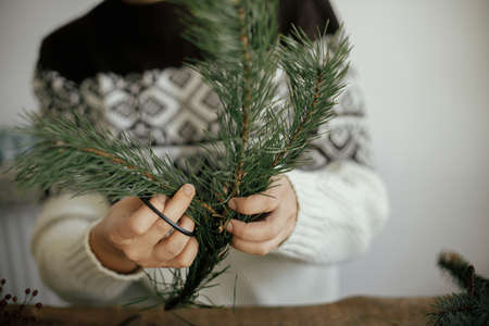Making christmas wreath. Woman in cozy sweater cutting pine branch with scissors on rustic wooden...