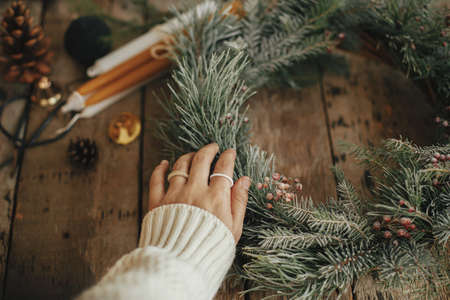 Hand with stylish rings on rustic christmas wreath with pine branches and red berries in snow on rustic old wooden background close up. Happy holidays. Making rustic xmas wreath, holiday workshopの写真素材