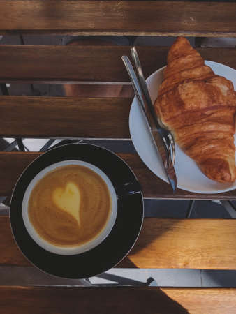 Delicious fresh croissant and coffee on wooden table in sunny light on cafe terrace. Top view. Fresh tasty breakfast in morning city. Aestheticsの写真素材