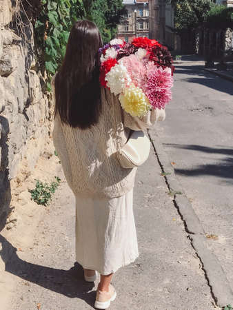 Stylish hipster woman with big autumn flowers bouquet walking in sunny european city street. Back view. Young brunette female in modern beige outfit walking with bunch of dahlias flowers. Aestheticsの写真素材