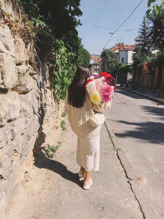 Stylish fashionable woman with big autumn flowers bouquet walking in sunny old street in european city. Young brunette female in modern beige outfit holding bunch of flowers. Aestheticsの写真素材