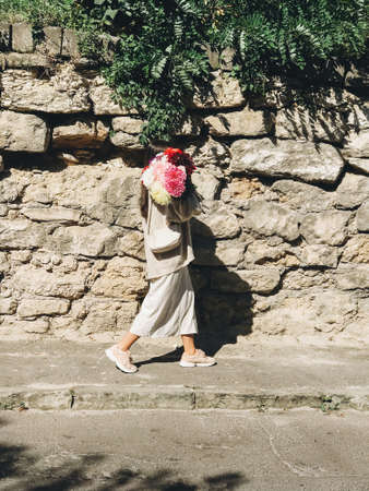 Stylish fashionable woman with big autumn flowers bouquet walking in sunny old street in european city. Young brunette female in modern beige outfit holding bunch of flowers. Aestheticsの写真素材