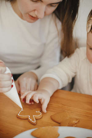 Cute little daughter and mother decorating christmas gingerbread cookies with icing on wooden table, close up. Family time together, xmas holiday preparations. Authentic momentの写真素材