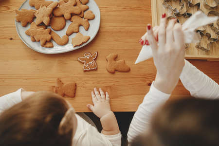 Cute little daughter and mother decorating christmas gingerbread cookies with icing on wooden table.Top view.  Family time, xmas holiday preparations. Mommy daughter authentic lovely momentsの写真素材