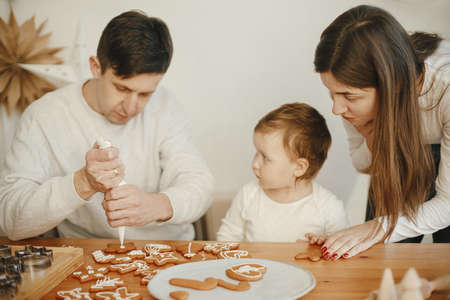 Stylish mother, father and cute little daughter decorating christmas gingerbread cookies with icing in modern room. Happy family time together, authentic lovely moments. Xmas holiday preparationsの写真素材