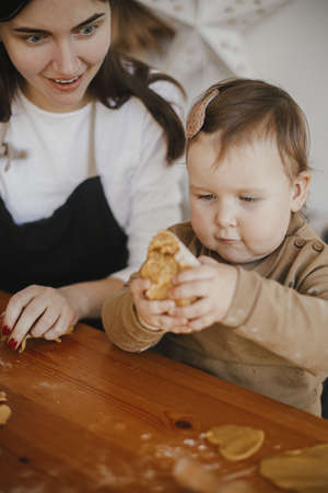 Adorable baby girl helping  mother kneading dough for gingerbread cookies on wooden table in modern...