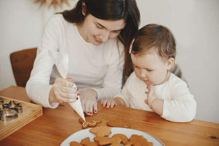 Cute little daughter and mother decorating christmas gingerbread cookies with frosting on wooden table in modern room. Happy family time together, holiday preparations. Happy Holidays!の写真素材