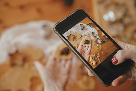 Woman making photo of gingerbread cookies for social media and blog. Hand holding phone and taking photo of heart shaped christmas cookies on messy table flat lay. Holiday preparationsの写真素材