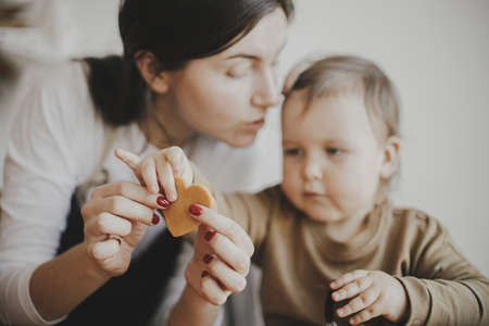 Adorable little daughter and mother making together gingerbread cookies on messy wooden table in...