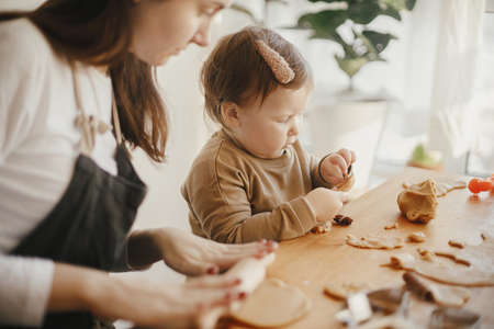 Adorable little daughter and mother making together gingerbread cookies on messy wooden table in modern room. Cute toddler girl helps making christmas cookies. Mommy daughter lovely momentsの写真素材