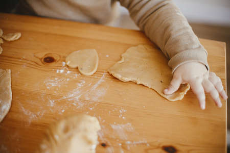 Adorable baby daughter kneading dough together with mother for gingerbread cookies on wooden table,...