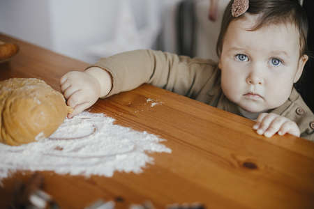 Adorable baby girl helping  mother kneading dough for gingerbread cookies on wooden table in modern scandinavian room. Cute toddler daughter helps making christmas cookies. Authentic momentの写真素材