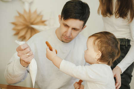 Stylish mother, father and cute little daughter decorating christmas gingerbread cookies with icing in modern room, funny authentic moments. Happy family time together. Xmas holiday preparationsの写真素材