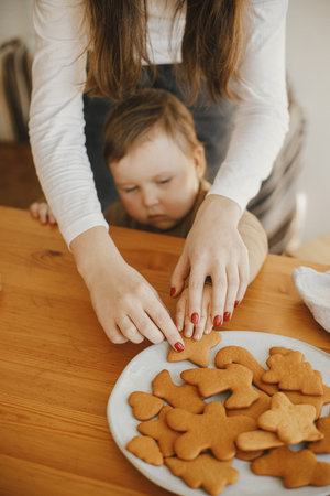 Cute little girl grabbing freshly baked gingerbread cookie on wooden table in modern room. Adorable toddler girl making christmas cookies with mother. Authentic lovely moment, holiday preparationの写真素材