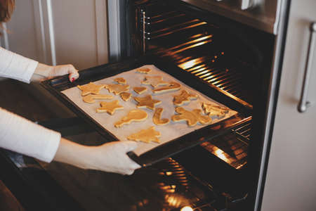 Woman putting tray with christmas cookies in oven close up in modern kitchen. Baking gingerbread cookies. Family holiday preparation, xmas culinary. Biscuits recipeの写真素材