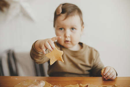 Cute little girl holding christmas star cookie on table, close up. Adorable toddler daughter helps mother making gingerbread cookies. Lovely moments, family time. Holiday preparationsの写真素材