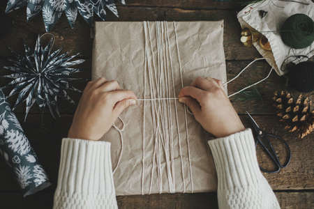 Hands wrapping stylish christmas gift in craft paper with string on rustic wooden table with blue paper stars. Flat lay. Atmospheric moody image, nordic style. Merry Christmas and Happy Holidays!の写真素材