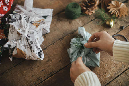 Christmas Furoshiki gift. Hands wrapping xmas gift in fabric on rustic wooden table with ornaments. Atmospheric moody image, nordic style. Zero waste and eco friendly presents. Holidays preparationの写真素材