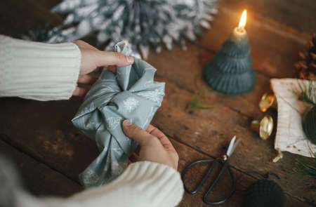 Hands wrapping christmas gift in fabric on rustic wooden table with candle, scissors, blue paper stars. Atmospheric moody image, nordic style. Merry Christmas and Happy Holidays! Furoshiki wrapの写真素材