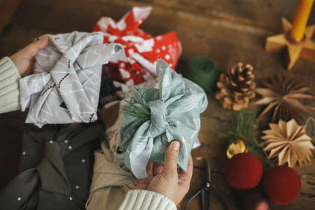 Hands holding christmas gift wrapped in fabric on rustic wooden table with ornaments. Atmospheric moody image, nordic style. Merry Christmas! Furoshiki wrap, zero waste holidaysの写真素材