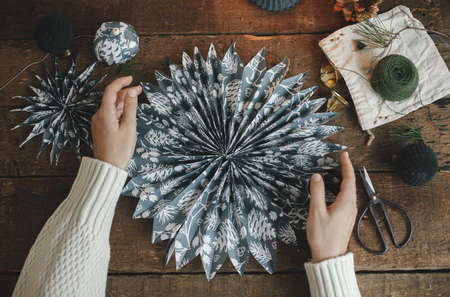 Hands holding stylish christmas star made of blue wrapping paper on rustic wooden table flat lay. Atmospheric moody image, nordic style. Handmade festive decor, holidays preparationの写真素材