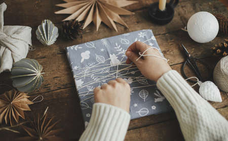 Hands wrapping christmas gift in fabric on rustic wooden table with scissors, craft paper star, candle. Atmospheric moody time, nordic style. Merry Christmas and Happy Holidays! Zero wasteの写真素材