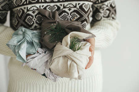 Woman hands in cozy sweater holding christmas gifts wrapped in fabric in scandinavian room. Atmospheric moody image, nordic style. Zero waste and eco friendly presents, Christmas Furoshiki giftsの写真素材