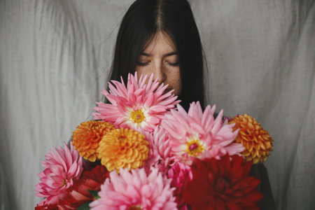 Beautiful woman smelling autumn flowers bouquet in rustic room. Atmospheric moody moment. Florist in linen dress holding beautiful colorful dahlias, aesthetic. Autumn season in countrysideの写真素材