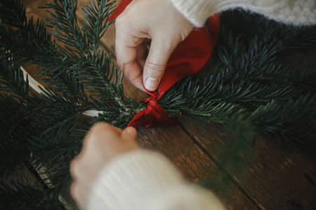 Hands making modern christmas wreath with fir branches and red ribbon on rustic table. Atmospheric moody image. Winter holidays preparation. Making minimalist xmas wreathの写真素材