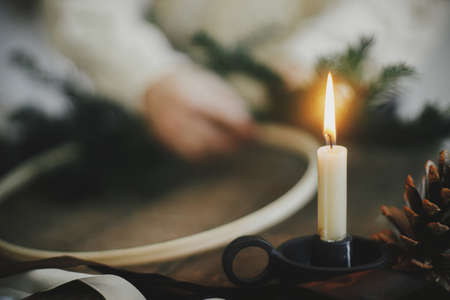 Stylish burning candle and pine cone on background of hands making modern christmas wreath with fir branches on rustic wooden table. Atmospheric image. Xmas advent. Winter holiday preparationの写真素材
