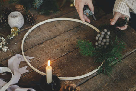 Making modern minimalistic christmas wreath. Hands making wreath on rustic table with fir branches, brunia herb, ribbons, round wooden hoop, thread, candle. Atmospheric moody imageの写真素材