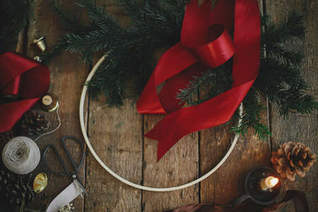 Making stylish modern christmas wreath flat lay. Fir branches, round wooden hoop, red ribbon, candle and pine cones on rustic table. Atmospheric moody image. Winter holidays preparationの写真素材