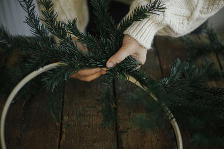 Hands making modern christmas wreath with fir branches on rustic table with round wooden hoop. Atmospheric moody image. Winter holidays preparation. Making minimalistic xmas wreathの写真素材