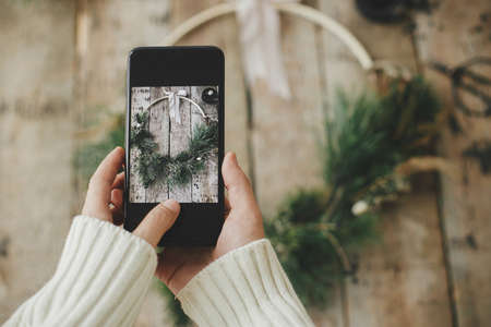 Hands holding phone and taking photo of modern christmas wreath rustic flat lay. Woman with smartphone making photo of stylish boho xmas wreath on rustic wood. Merry Christmas! Moody imageの写真素材