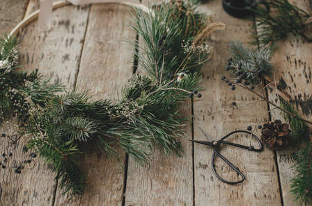Modern christmas wreath with spruce branches, herbs, scissors, pine cones on rustic wood. Merry Christmas! Stylish boho xmas wreath details. Moody image, winter holiday preparationの写真素材