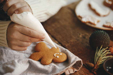 Hands decorating christmas gingerbread man cookie with frosting on rustic table with napkin, spices,...