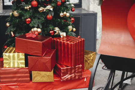 Stylish christmas red and gold wrapped gift boxes under modern traditional xmas tree with baubles outdoor. Christmas festive street decor for winter holidays in city street. Merry Christmas!の写真素材