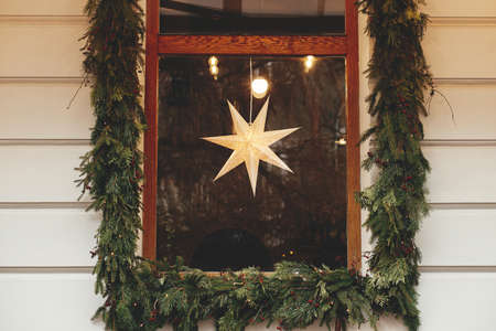 Stylish modern christmas star hanging in window with spruce branches on old building or shop. Merry Christmas. Illuminated sweden star. Christmas festive decor for winter holidaysの写真素材