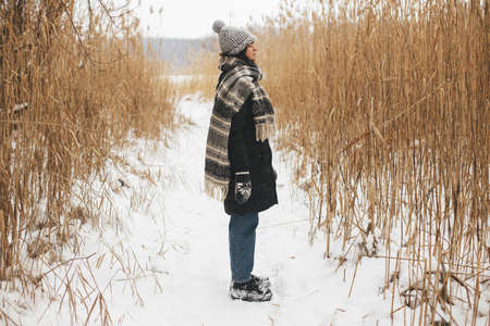 Stylish hipster woman standing among dried grass at snowy winter lake. Atmospheric calm moment. Tranquility. Young female in cozy stylish clothes relaxing in snowy park. Wanderlustの写真素材