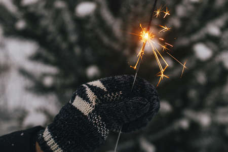 Hand in cozy mitten holding burning sparkler on background of pine tree branches in snow. Happy New Year! Atmospheric magic moment. Woman hand with glowing firework in evening.の写真素材