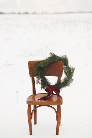 Merry Christmas! Christmas wreath on rustic chair in snowy winter field. Winter holidays in countryside. Space for text. Stylish xmas wreath with pine branches and red bow hanging on wooden chairの写真素材