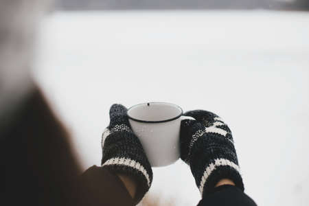 Hands in cozy gloves holding warm cup of tea on background of  snow lake in winter. Hiking and...