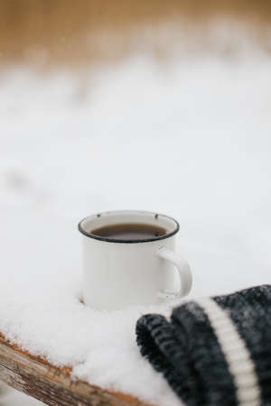 Warm cup of tea and cozy knitted gloves in snow on wooden bench in winter park. Hiking and traveling in cold season. Wanderlust. Space for text. Winter timeの写真素材