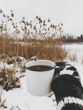 Hand in cozy glove holding warm cup of tea on background of dried grass and snow lake in winter....