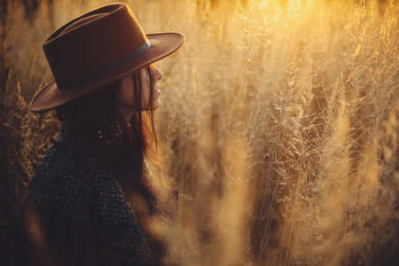 Portrait of beautiful stylish woman in hat standing in grass in warm sunset light in autumn field. Tranquil atmospheric moment. Fashionable young female enjoying evening in countrysideの写真素材
