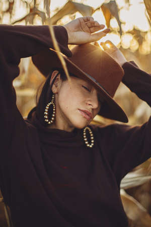 Portrait of stylish woman in hat and brown clothes posing in autumn maize field in sunset sunlight. Fashionable attractive young female  relaxing in autumnal corn in evening countrysideの写真素材