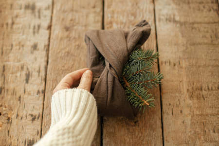Hand holding stylish christmas gift wrapped in fabric with fir branch on rustic wooden table. Zero waste winter holidays and gift. Furoshiki eco friendly present. Happy holidays and Merry Christmasの写真素材