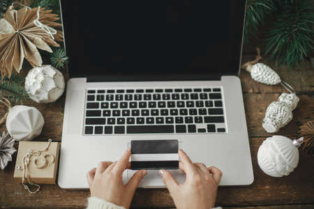 Christmas sale and shopping online. Hands with credit card on laptop with black screen on background of rustic wooden table with stylish christmas ornaments. View above. Space for texの写真素材