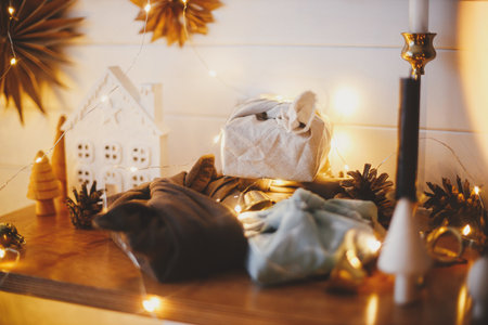 Stylish zero waste christmas gifts on wooden shelf on background of little house, trees, paper stars and christmas golden lights bokeh. Festive decorated scandinavian room. Atmospheric eveの写真素材
